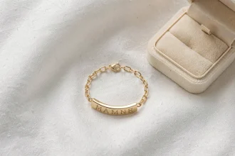 Gourmette en or gravée pour bébé posée sur tissu blanc avec écrin ouvert, symbole d'un bijou de baptême précieux à assurer contre la perte et le vol